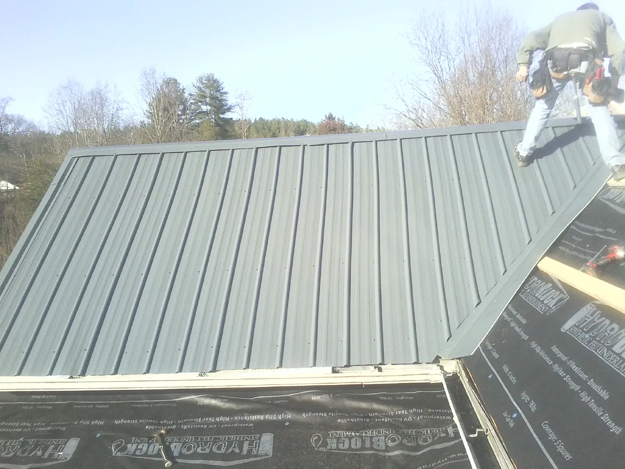 Crew installing dark green standing seam panels over synthetic underlayment in Grain Valley