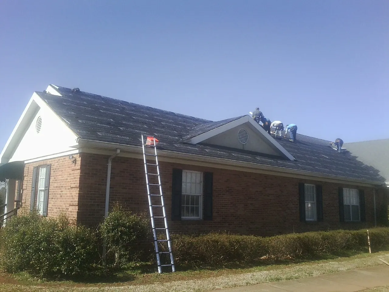 Crew tearing shingles off a brick ranch roof with a ladder set — Metal Roof Repair work by our crew in Grain Valley