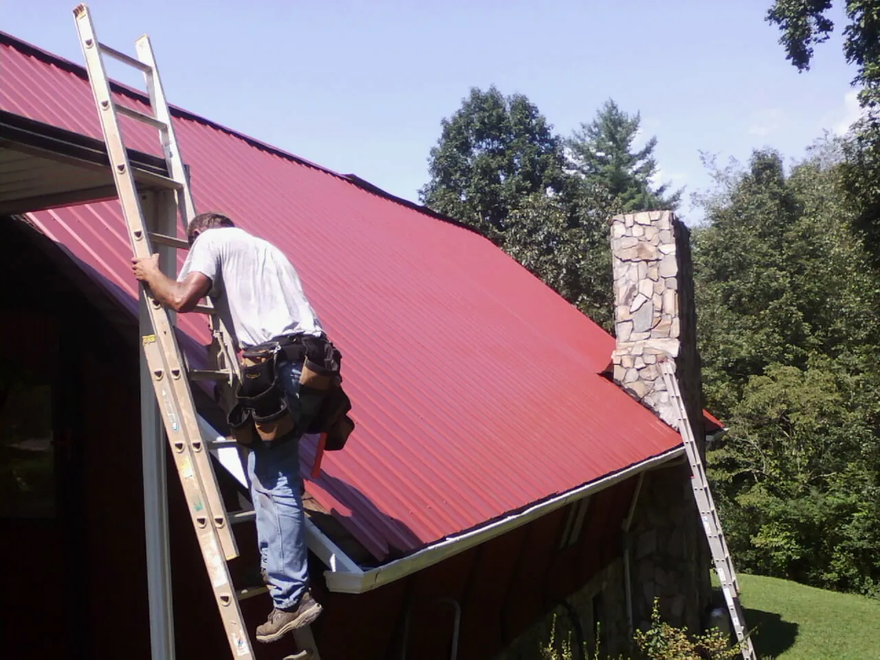 Roofer on a ladder beside a red metal roof and stone chimney — Soffit & Fascia Repair by Roofing Brothers in Grain Valley