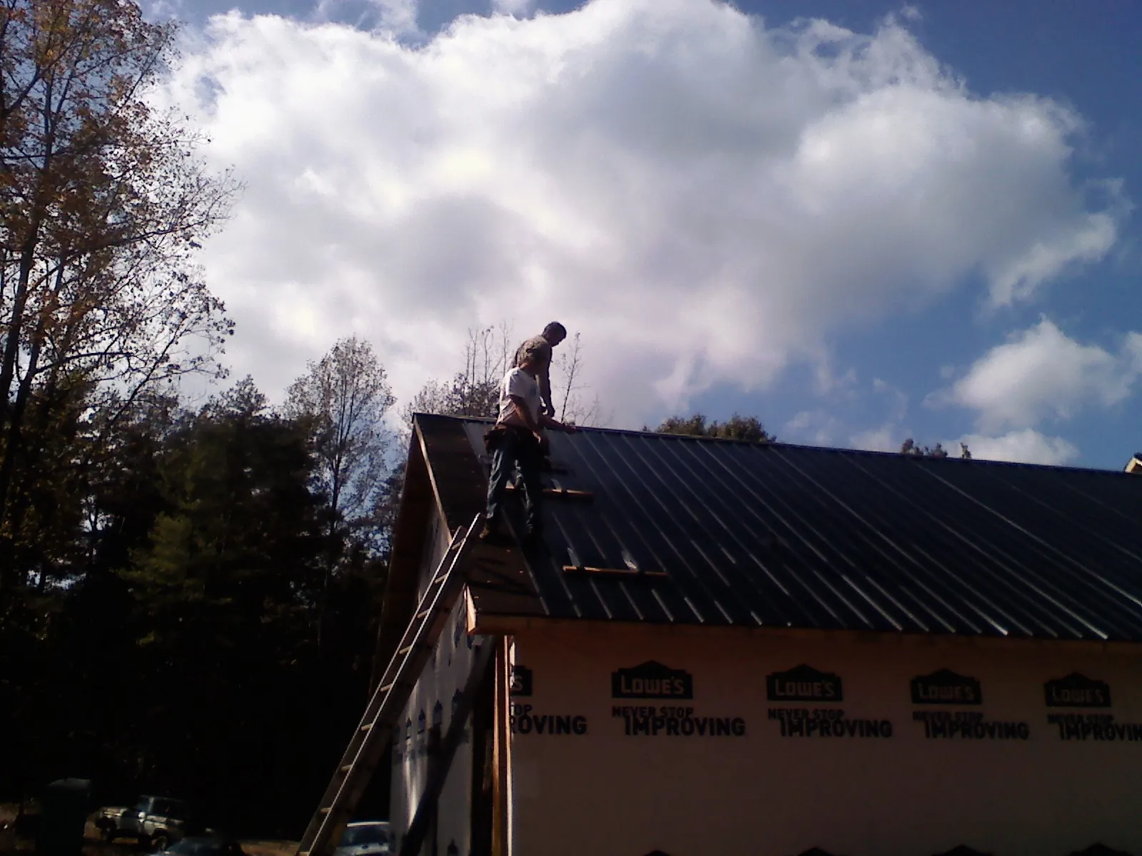 Roofer fastening dark metal panels high against a cloudy sky, from a Roof Inspection job in Grain Valley